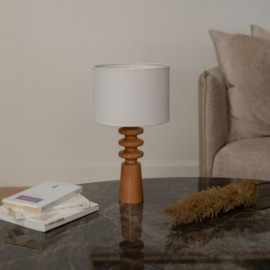 Verschönern Sie Ihren Wohnraum mit dieser eleganten Tischleuchte aus Holz mit einem skulpturalen Sockel und einem klassischen Lampenschirm aus Stoff. Die warmen Holztöne in Kombination mit dem minimalistischen Design machen sie zu einer vielseitigen Ergänzung für jedes Interieur, von modern bis rustikal. Diese Lampe eignet sich perfekt für Nachttische, Wohnzimmer oder Heimbüros und sorgt für ein sanftes Umgebungslicht, das eine gemütliche Atmosphäre schafft. Der handgefertigte Holzsockel verleiht ihr einen 